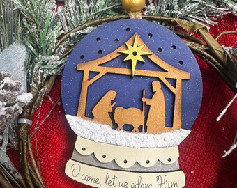 May include: A Christmas ornament shaped like a snow globe, featuring a nativity scene. The ornament has a blue background with a gold star and a wooden stable. The text "O come, let us adore Him" is written on the bottom.