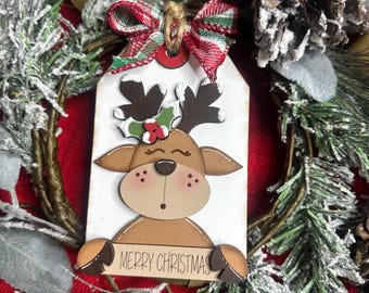 May include: A wooden Christmas ornament featuring a reindeer design with holly accents and the text "MERRY CHRISTMAS." The ornament is attached to a red and green plaid bow and a jute string, surrounded by festive greenery and red fabric.