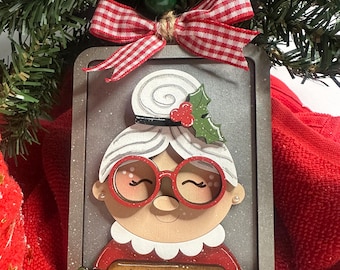 May include: A rectangular Christmas ornament featuring a cartoon depiction of a woman with glasses, a white hair bun, and a red outfit. The ornament has the word "Nana" on a rolling pin. It is decorated with a red and white checkered bow and a green bead.