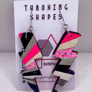 May include: Handmade lightning bolt earrings in black, silver, white, and neon pink leather. The earrings are attached to a backing card with the text "THROWING SHAPES". The design is geometric and bold.
