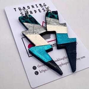 May include: Handmade lightning bolt earrings in teal, silver, white, black, and teal leopard print. The earrings are layered and hang from silver hooks. The earrings are displayed on a card with the text "THROWING SHAPES".