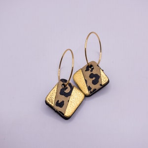 May include: A pair of gold hoop earrings with square-shaped dangles. The dangles are made of brown leather with a black leopard print and gold accents.