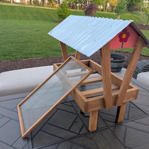 May include: A wooden bird feeder with a corrugated metal roof and a red flower design. The feeder has a slanted glass panel and is supported by wooden posts. The feeder is on a dark gray table.