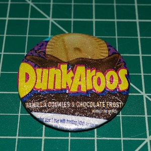 May include: A round, colorful button with the words "Dunkaroos" in yellow letters on a purple background. The button also features a picture of a cookie with chocolate frosting and the text "Vanilla Cookies & Chocolate Frosting".
