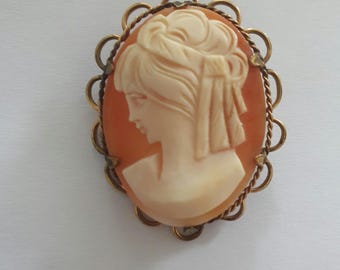 May include: A cameo brooch featuring a carved portrait of a woman with a detailed hairstyle. The cameo is set in a gold-toned metal frame with a decorative rope-like design.