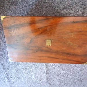 May include: A rectangular wooden box with a polished brown grain finish, featuring brass corner protectors and a central brass inlay shield. The lid is smooth with visible wood grain patterns and light surface scratches across the top surface.