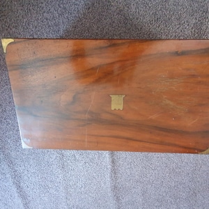 Può includere: Una scatola di legno a forma di trapezio, di colore marrone intenso con motivi di venature più scure. La scatola ha angoli in ottone e un piccolo emblema in ottone al centro. Il design suggerisce uno stile vintage o antico.