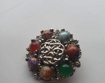 Può includere: Un fermaglio in argento con un nodo celtico a forma circolare decorato con sette pietre colorate. Le pietre sono rosse, arancioni, marroni, verdi, blu, viola e bianche.