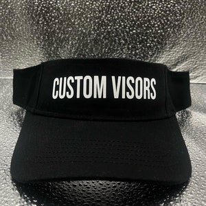 May include: A black visor featuring the text CUSTOM VISORS in white capital letters printed on the front panel. The visor has a curved brim and a structured crown design, constructed from a solid black fabric with visible stitching details.