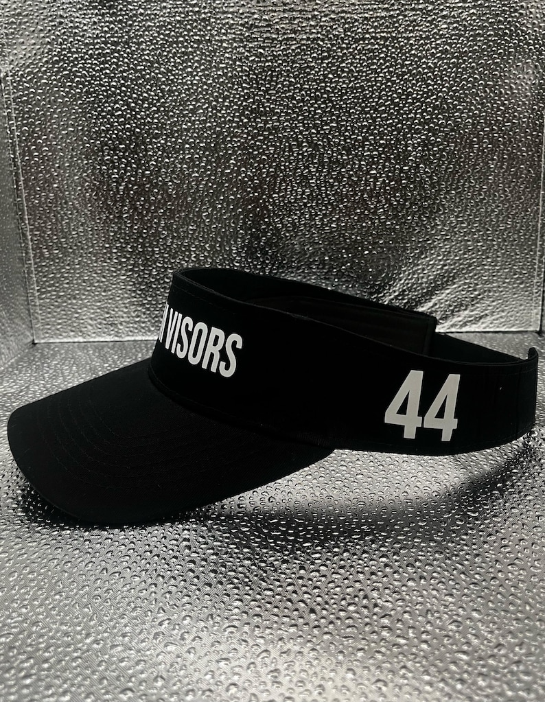 May include: A black visor featuring the white text VISORS printed on the front panel and the number 44 in white on the side. The headwear has a curved brim and an adjustable strap at the back, constructed from a solid black fabric material.