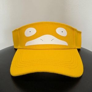 May include: A bright yellow visor featuring a white graphic print of a stylized duck-like face with two circular eyes and a wide, flat beak centered on the front panel. The visor has a curved brim and a structured crown made of fabric.