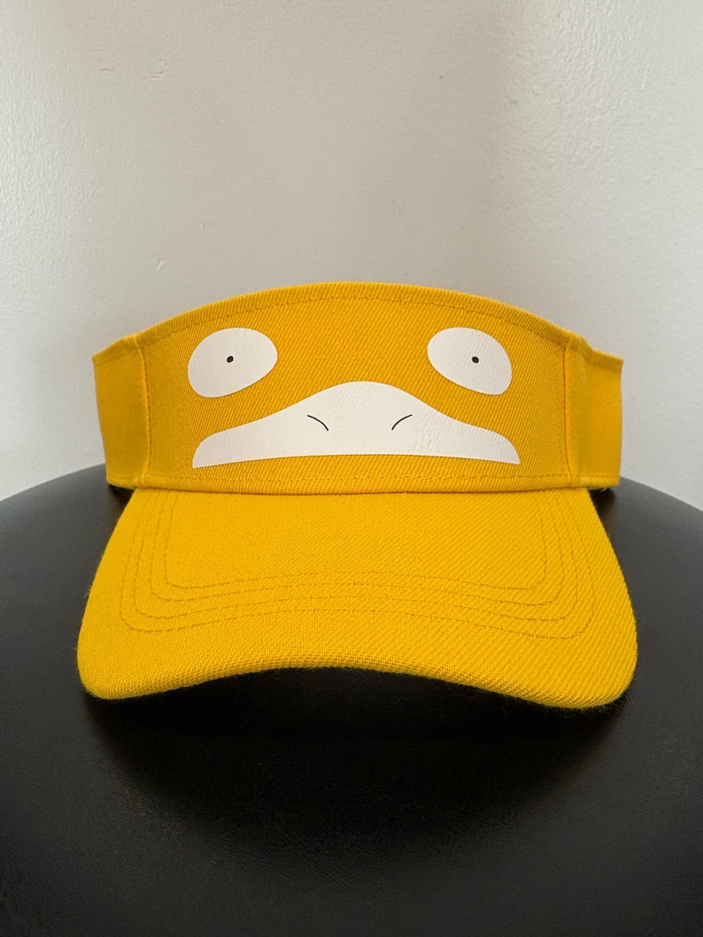 May include: A bright yellow visor featuring a white graphic print of a stylized duck-like face with two circular eyes and a wide, flat beak centered on the front panel. The visor has a curved brim and a structured crown made of fabric.