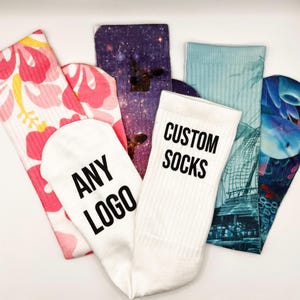 以下が含まれることがあります： 異なるデザインのカラフルな靴下5足セット。1足は「CUSTOM SOCKS」という文字が印刷された白い靴下。もう1足は「ANY LOGO」という文字が印刷された白い靴下。他の靴下は、銀河柄、花柄、青と白の海柄、青と白の船柄です。