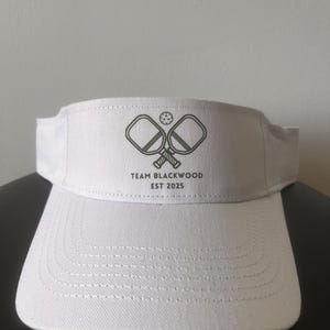 May include: A white visor featuring a dark gray graphic of two crossed pickleball paddles and a ball. Below the graphic, the text reads "TEAM BLACKWOOD" and "EST 2025". The visor has a structured front panel with visible stitching on the brim.