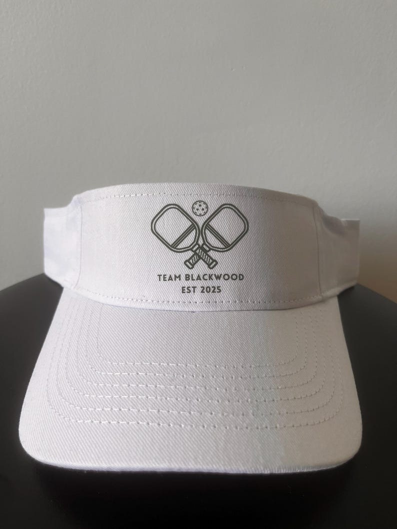 May include: A white visor featuring a dark gray graphic of two crossed pickleball paddles and a ball. Below the graphic, the text reads "TEAM BLACKWOOD" and "EST 2025". The visor has a structured front panel with visible stitching on the brim.