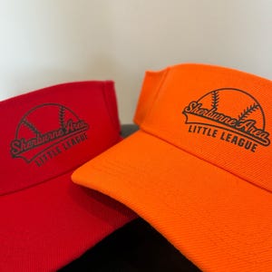 May include: A red visor and an orange visor, each featuring a black graphic of a baseball with the text Sherburne Area LITTLE LEAGUE printed across the front. Both visors have a curved brim and a structured crown design for athletic wear.