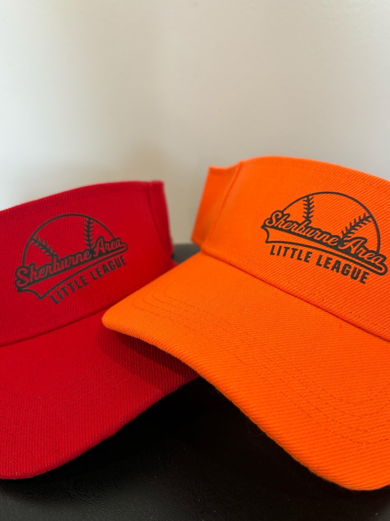 May include: A red visor and an orange visor, each featuring a black graphic of a baseball with the text Sherburne Area LITTLE LEAGUE printed across the front. Both visors have a curved brim and a structured crown design for athletic wear.