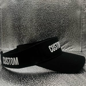 May include: A black visor featuring the word CUSTOM printed in white capital letters on both the front brim and the side band. The visor has a curved brim and an adjustable strap at the back, constructed from a solid black fabric material.