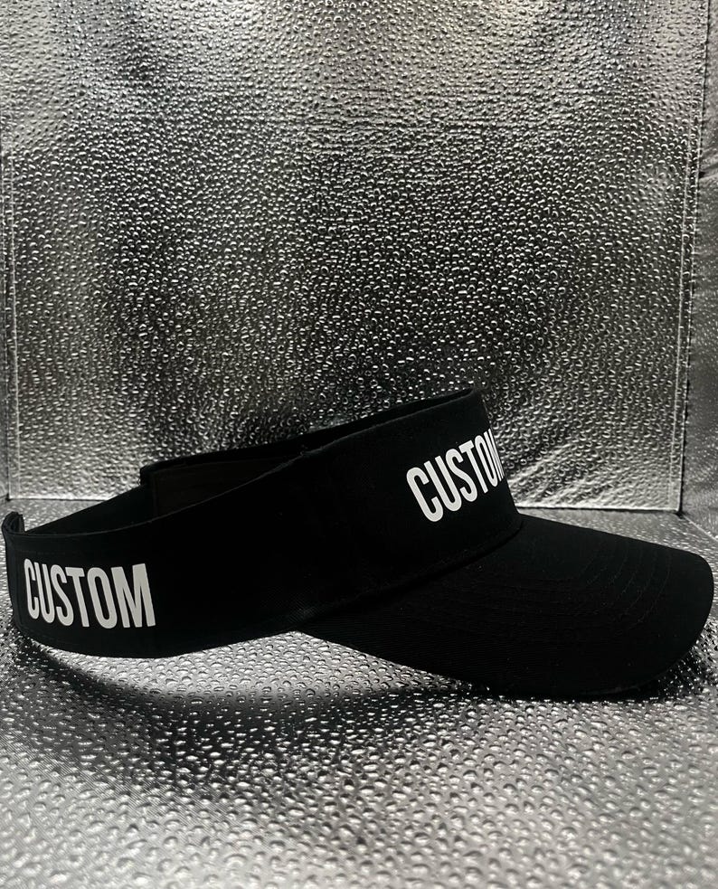 May include: A black visor featuring the word CUSTOM printed in white capital letters on both the front brim and the side band. The visor has a curved brim and an adjustable strap at the back, constructed from a solid black fabric material.