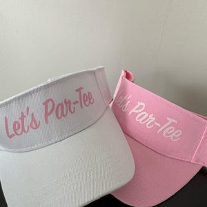 May include: Two sun visors, one white and one light pink, both featuring the text Let's Par-Tee embroidered in a script font on the front panel. The visors have a structured brim and an adjustable strap design, showcasing a clean, simple aesthetic.