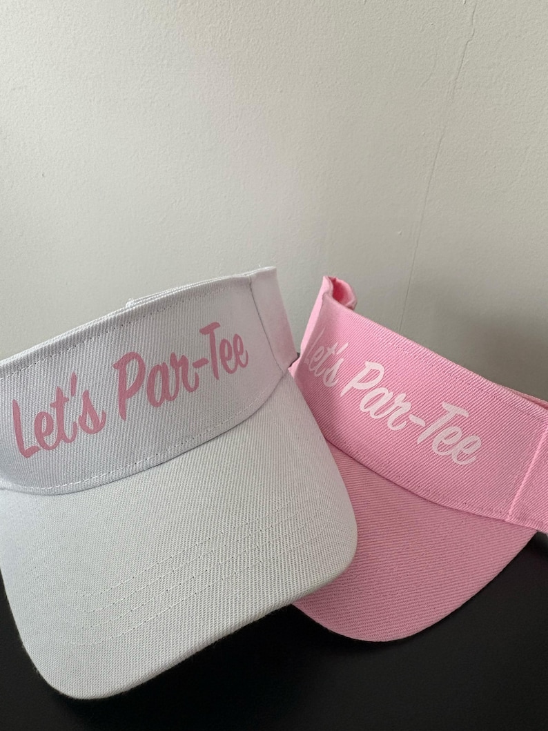 May include: Two sun visors, one white and one light pink, both featuring the text Let's Par-Tee embroidered in a script font on the front panel. The visors have a structured brim and an adjustable strap design, showcasing a clean, simple aesthetic.