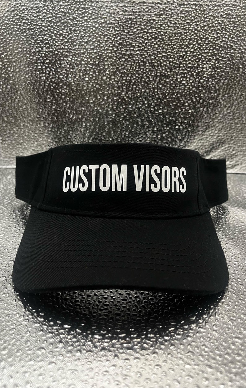 May include: A black visor featuring the text CUSTOM VISORS in white capital letters printed on the front panel. The visor has a curved brim and a structured crown design, constructed from a solid black fabric with visible stitching details.