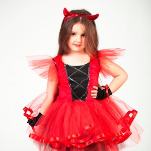 May include: A young girl dressed as a devil for Halloween. She is wearing a red tulle dress with black trim and red ribbon accents. She has a red devil horn headband on her head and black gloves on her hands.