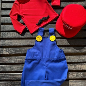 Puede incluir: Una camisa roja de manga larga con un bigote negro de aplique, un mono azul con botones amarillos y un gorro rojo. Este es un disfraz para ni&ntilde;os.