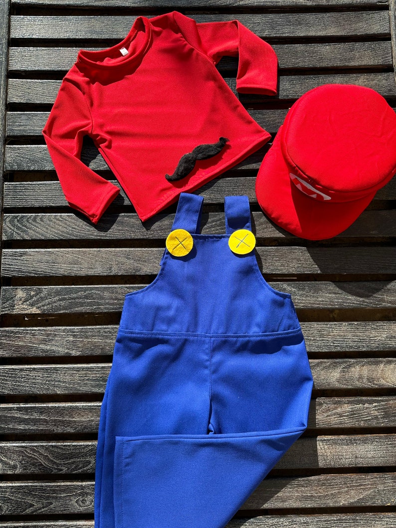 Puede incluir: Una camisa roja de manga larga con un bigote negro de aplique, un mono azul con botones amarillos y un gorro rojo. Este es un disfraz para ni&ntilde;os.