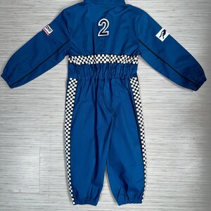 May include: Blue racing jumpsuit with black and white checkered accents. The jumpsuit has a zipper closure, a collar, and long sleeves. The back of the jumpsuit has a number "2" and a kangaroo logo.