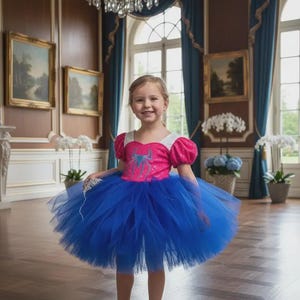 May include: A young girl wearing a vibrant blue tutu dress with a pink bodice featuring a spider graphic. The dress has short, puffy sleeves and is paired with blue shoes. The background includes a chandelier and ornate interior.