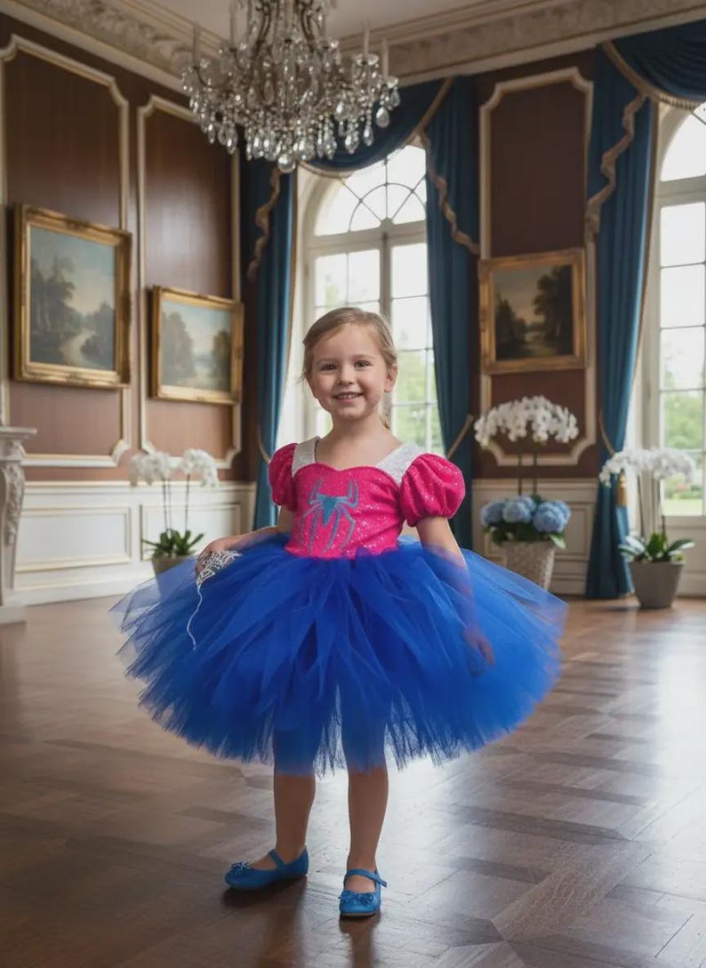 May include: A young girl wearing a vibrant blue tutu dress with a pink bodice featuring a spider graphic. The dress has short, puffy sleeves and is paired with blue shoes. The background includes a chandelier and ornate interior.