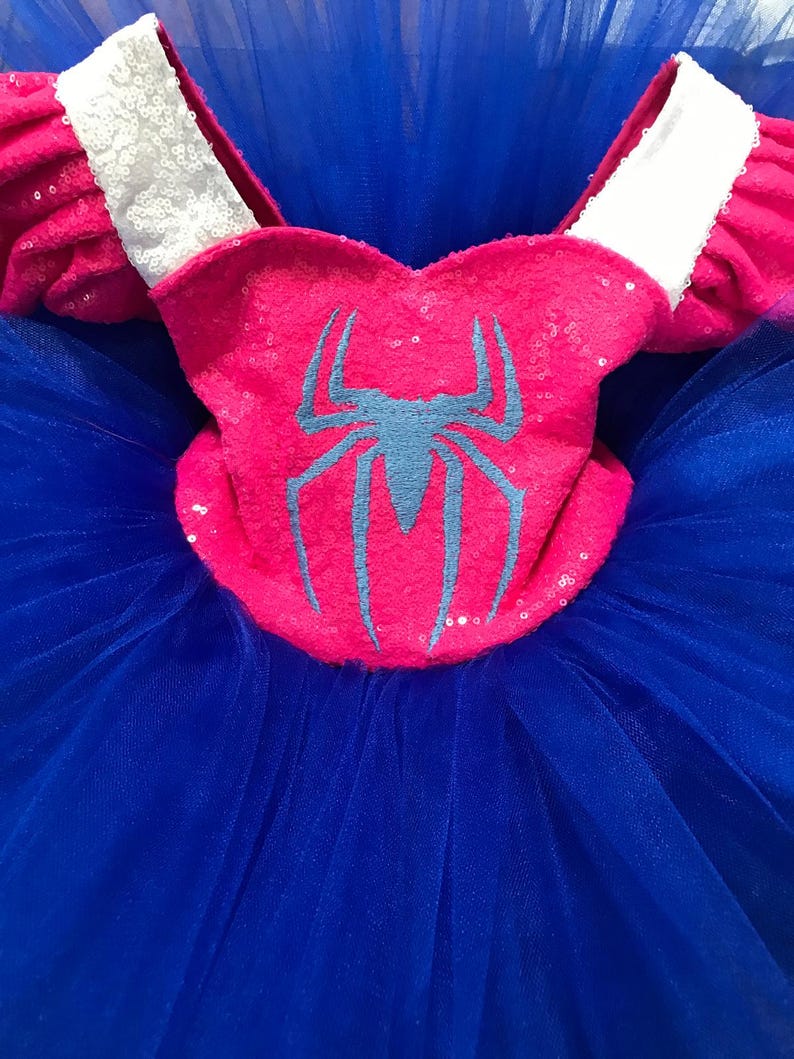 May include: A vibrant pink and blue costume dress. The bodice is pink with a blue spider emblem and silver sequins. The sleeves are pink with white sequined trim. The skirt is made of blue tulle.
