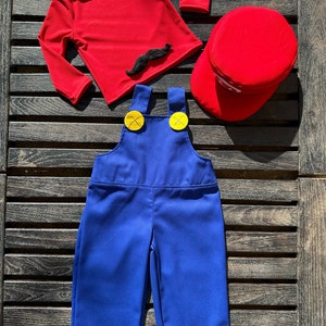 Puede incluir: Una camisa roja de manga larga con un bigote negro de aplique, un mono azul con botones amarillos y una gorra roja. Este es un disfraz para ni&ntilde;os.