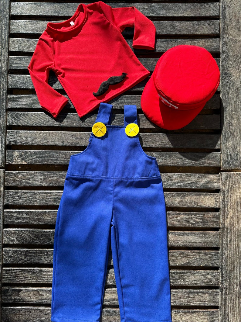 Puede incluir: Una camisa roja de manga larga con un bigote negro de aplique, un mono azul con botones amarillos y una gorra roja. Este es un disfraz para ni&ntilde;os.