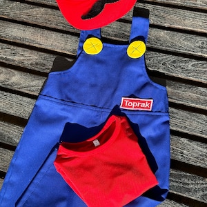 Puede incluir: Un mono azul con botones amarillos y una camiseta roja. El mono tiene un parche que dice "Toprak". Un sombrero rojo con una "M" blanca y un bigote negro est&aacute; encima del mono.