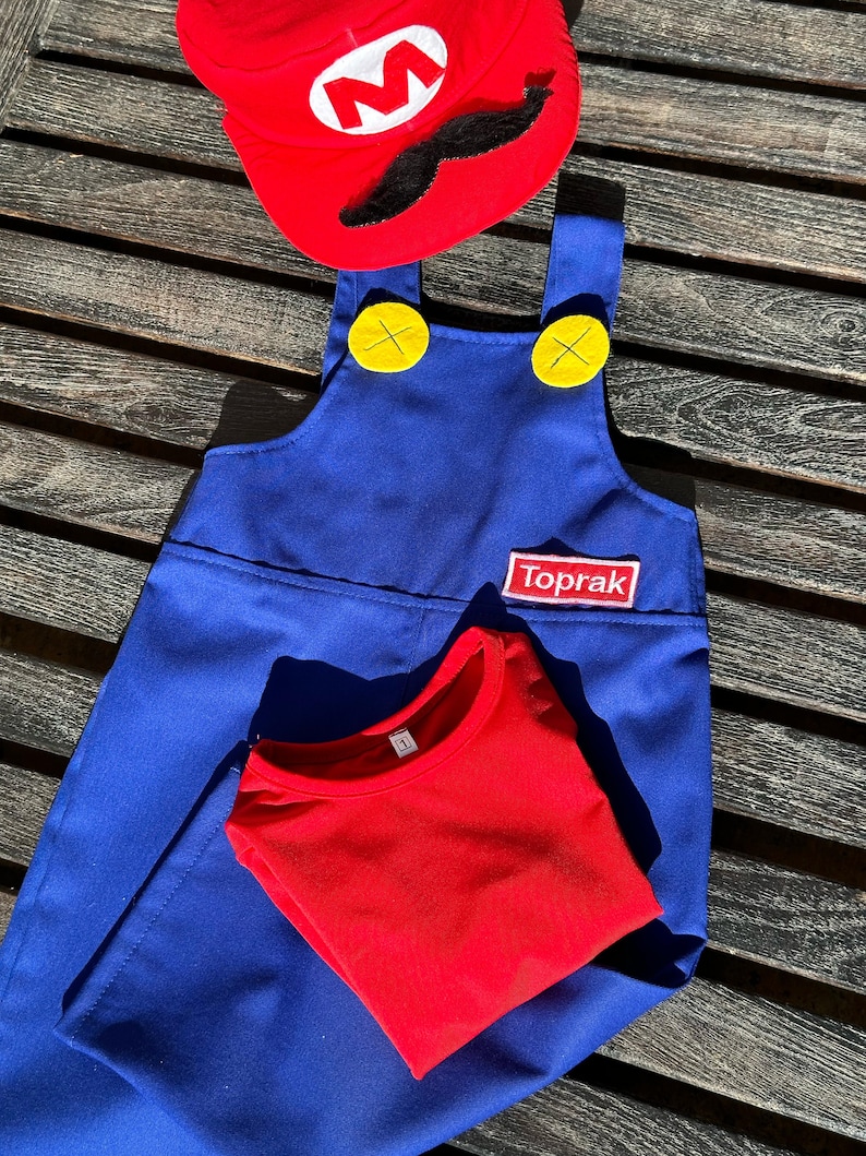 Puede incluir: Un mono azul con botones amarillos y una camiseta roja. El mono tiene un parche que dice "Toprak". Un sombrero rojo con una "M" blanca y un bigote negro est&aacute; encima del mono.