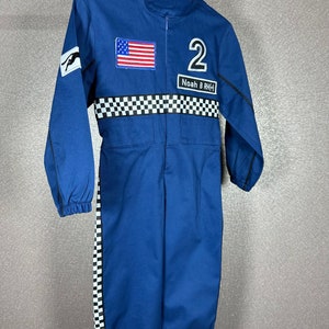 May include: Blue racing jumpsuit with a checkered pattern on the sleeves and legs. The jumpsuit has a zipper closure and a patch with the American flag on the chest. The number "2" and the name "Noah B. RHCK" are embroidered on the front.