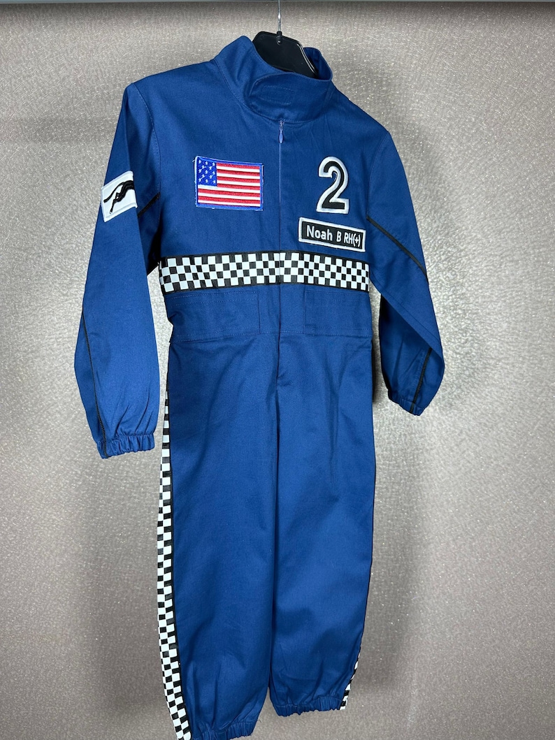 May include: Blue racing jumpsuit with a checkered pattern on the sleeves and legs. The jumpsuit has a zipper closure and a patch with the American flag on the chest. The number "2" and the name "Noah B. RHCK" are embroidered on the front.