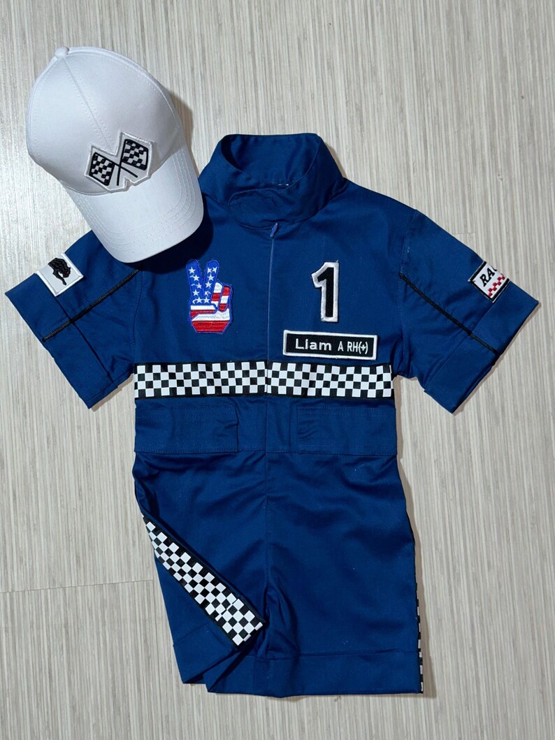 May include: A blue racing jumpsuit with a checkered pattern, a peace sign with an American flag, and the number 1. The jumpsuit has the text "Liam A RH(+) ". A white baseball cap with a checkered flag design is also included.