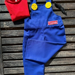 Puede incluir: Pantal&oacute;n vaquero azul con botones amarillos y una camiseta roja. El pantal&oacute;n vaquero tiene un parche que dice "Toprak" en la parte delantera.