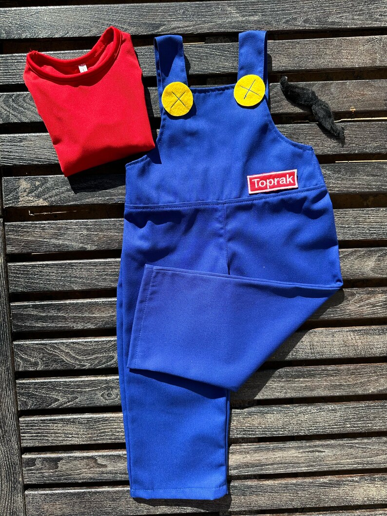 Puede incluir: Pantal&oacute;n vaquero azul con botones amarillos y una camiseta roja. El pantal&oacute;n vaquero tiene un parche que dice "Toprak" en la parte delantera.