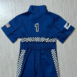 May include: A blue racing jumpsuit with a white checkered pattern on the sleeves and waist. The jumpsuit has a black number "1" on the back and a black and white logo on the right sleeve.