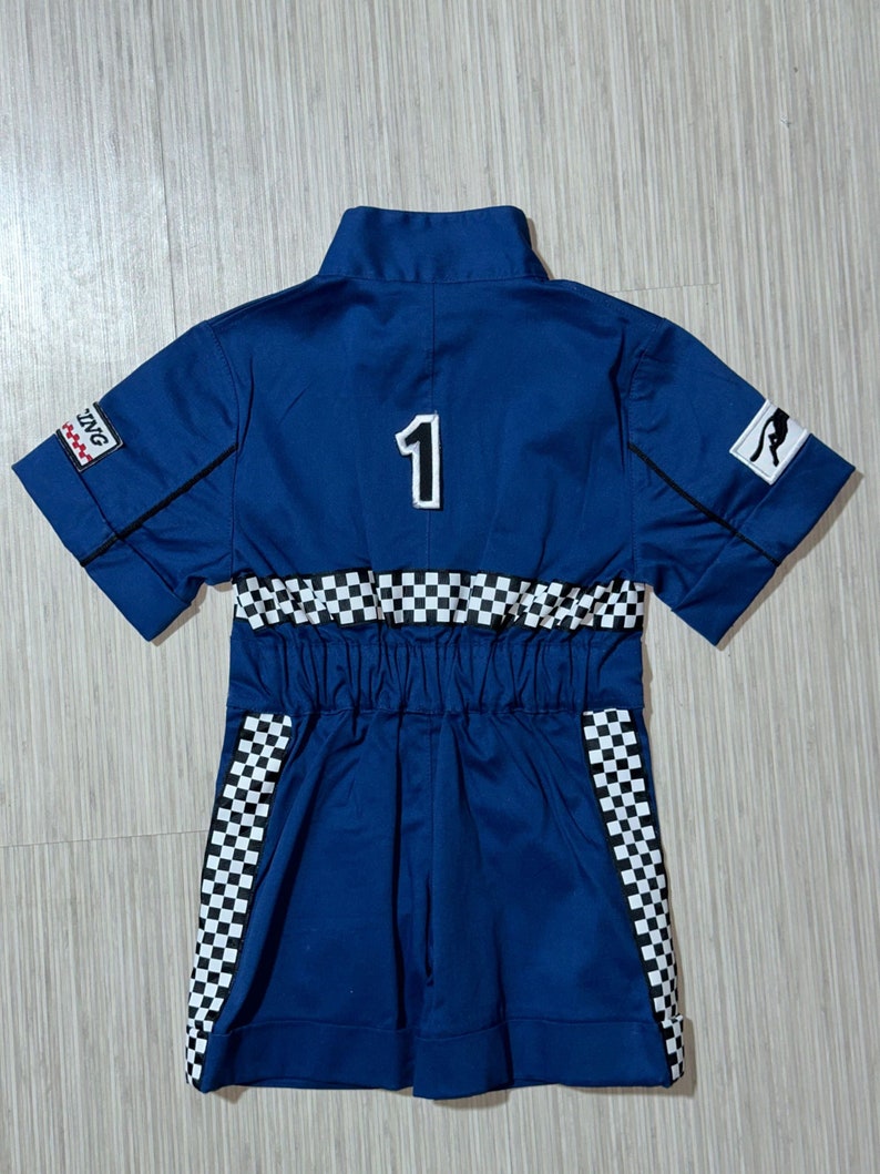 May include: A blue racing jumpsuit with a white checkered pattern on the sleeves and waist. The jumpsuit has a black number "1" on the back and a black and white logo on the right sleeve.