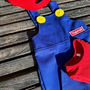 Puede incluir: Un mono azul con botones amarillos y un parche rojo que dice "Toprak". El mono est&aacute; sobre una superficie de madera con un sombrero rojo con una "M" blanca y un bigote negro en la parte superior.