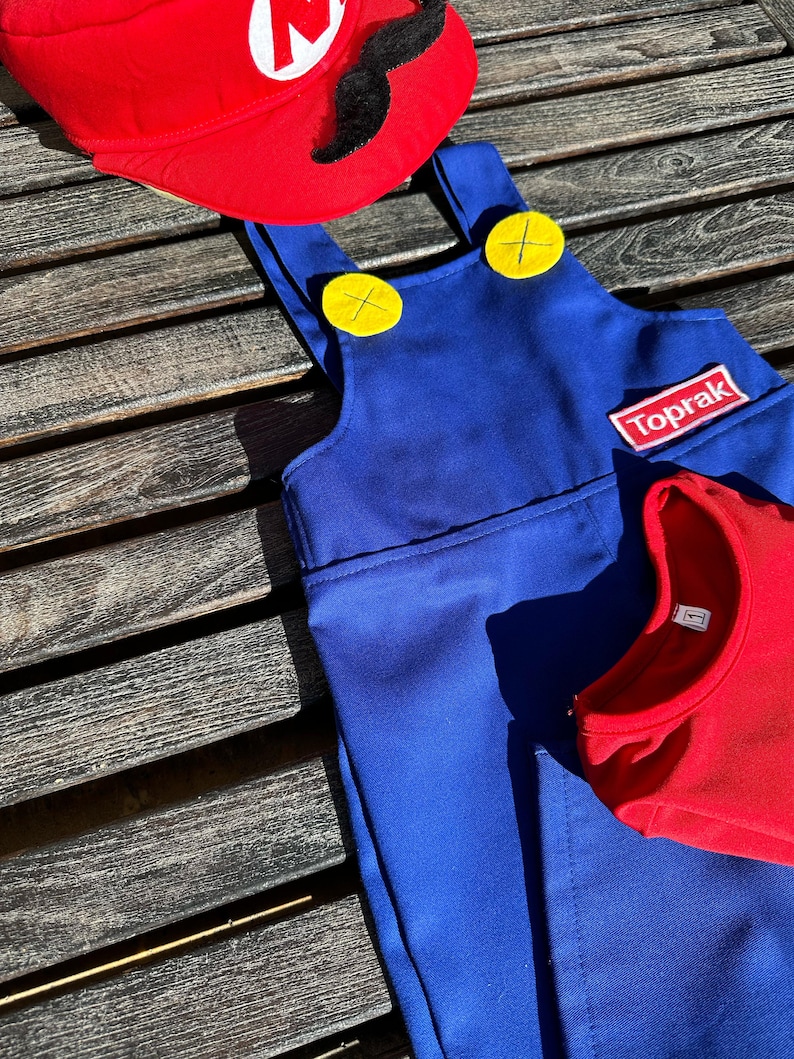 Puede incluir: Un mono azul con botones amarillos y un parche rojo que dice "Toprak". El mono est&aacute; sobre una superficie de madera con un sombrero rojo con una "M" blanca y un bigote negro en la parte superior.