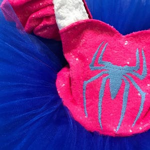 May include: A close-up of a pink sequined accessory with a blue spider emblem, set against a backdrop of blue tulle. The accessory has a white sequined trim. The image showcases a costume element, likely for a child's outfit.