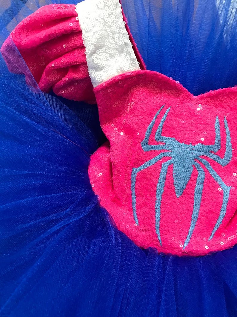 May include: A close-up of a pink sequined accessory with a blue spider emblem, set against a backdrop of blue tulle. The accessory has a white sequined trim. The image showcases a costume element, likely for a child's outfit.