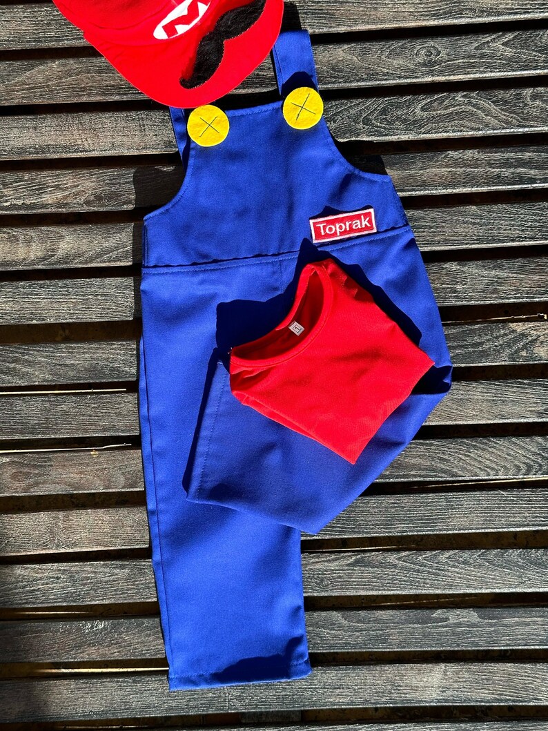 Puede incluir: Pantal&oacute;n vaquero azul con botones amarillos y una camiseta roja. El pantal&oacute;n vaquero tiene un parche que dice "Toprak" en la parte delantera.
