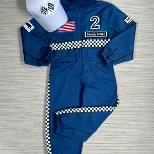 May include: A blue racing jumpsuit with black and white checkered trim. The jumpsuit has a number "2" and the name "Noah B (RH)" on the front. It also has an American flag patch and a black and white checkered flag patch. A white baseball cap with a black and white checkered flag patch is included.